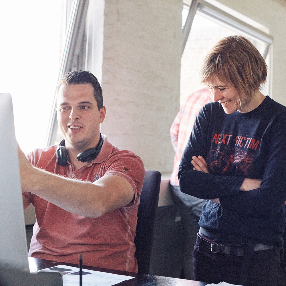 Man sieht zwei Mitarbeiter von FederhenSchneider, eine Frau und einen Mann, vor einem Monitor, die lachen.