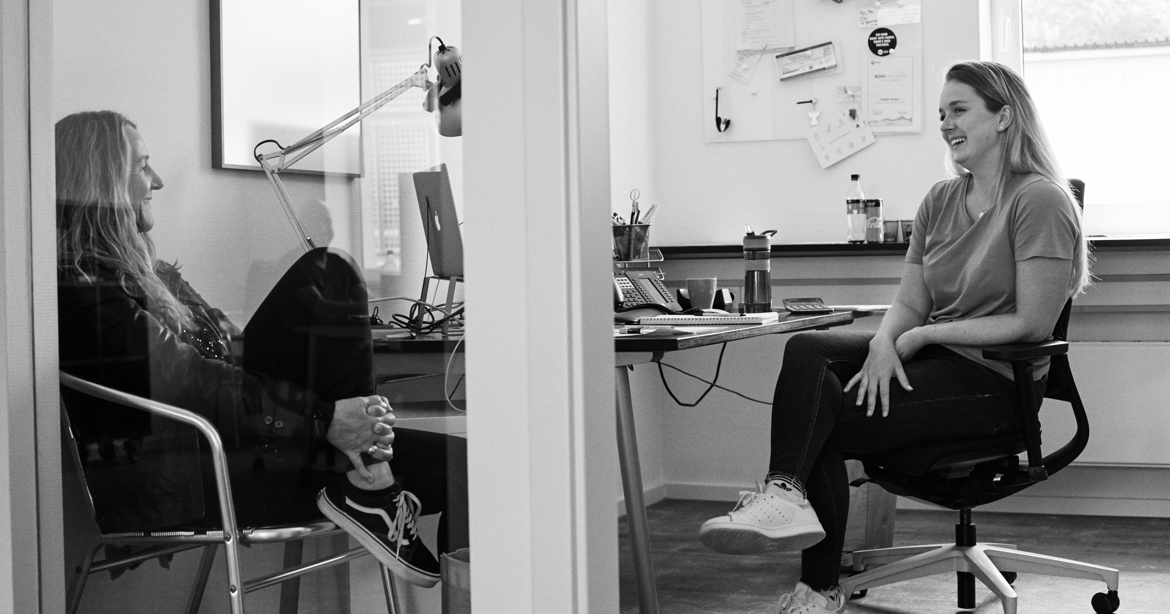 Man sieht zwei Mitarbeiterinnen von FederhenSchneider, die in einem Büro am Schreibtisch sitzen und sich unterhalten. Das Bild ist schwarz/weiß.