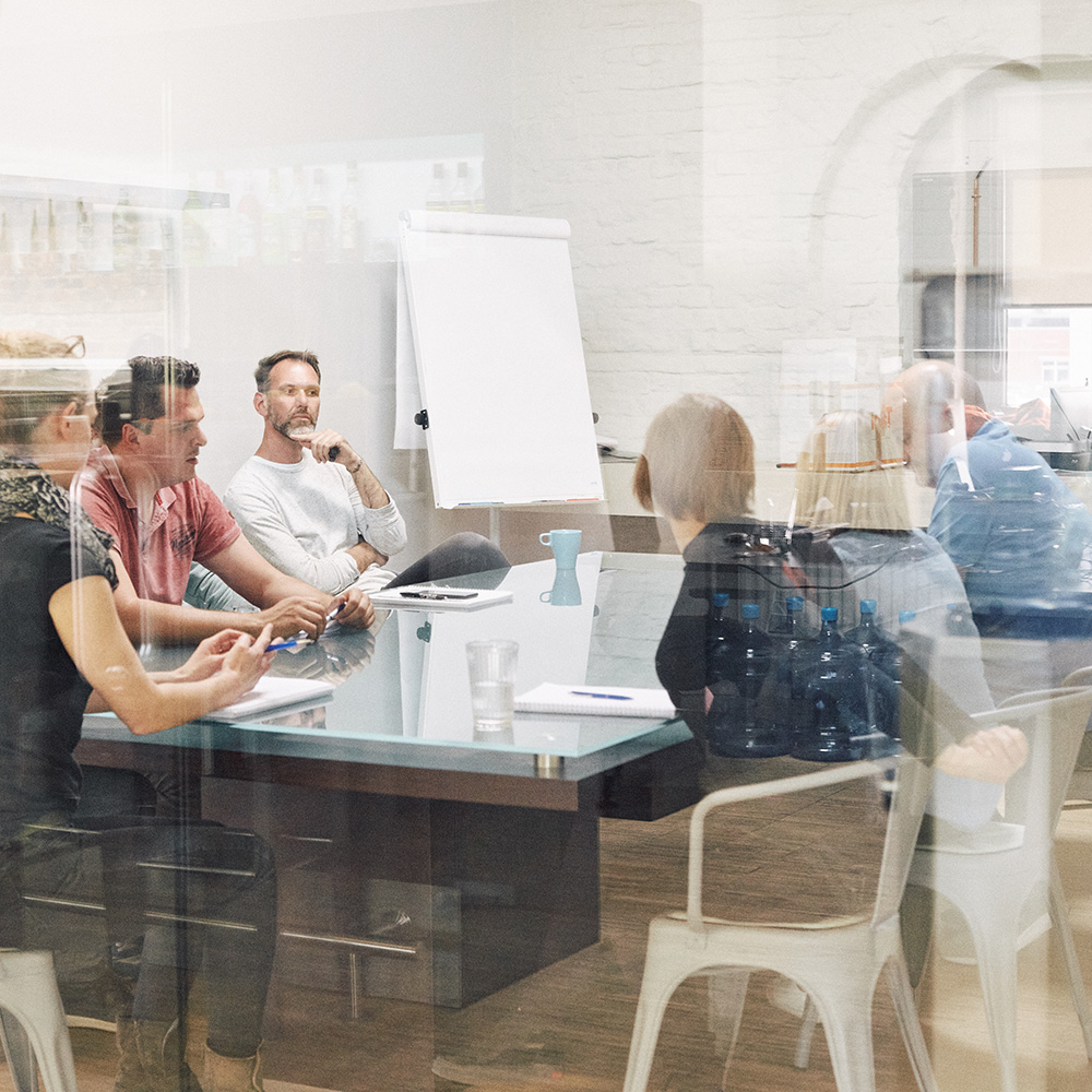 Man sieht mehrere Mitarbeiter von FederhenSchneider unterschiedlichen Geschlechts und Alters in einem Konferenzraum durch die Scheibe fotografiert. Sie sehen nachdenklich aus.