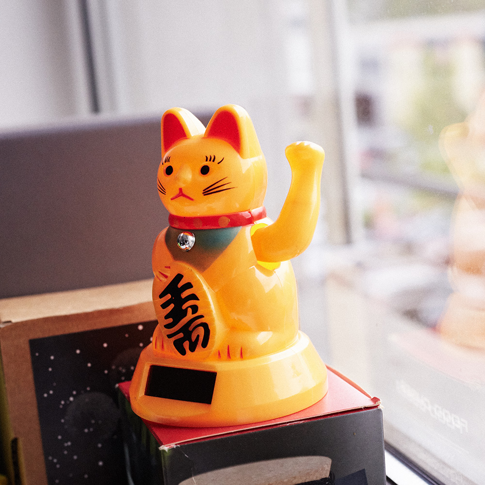 Das Bild zeigt eine orangene Winke-Katze aus Plastik, die ein einem Fenster steht.