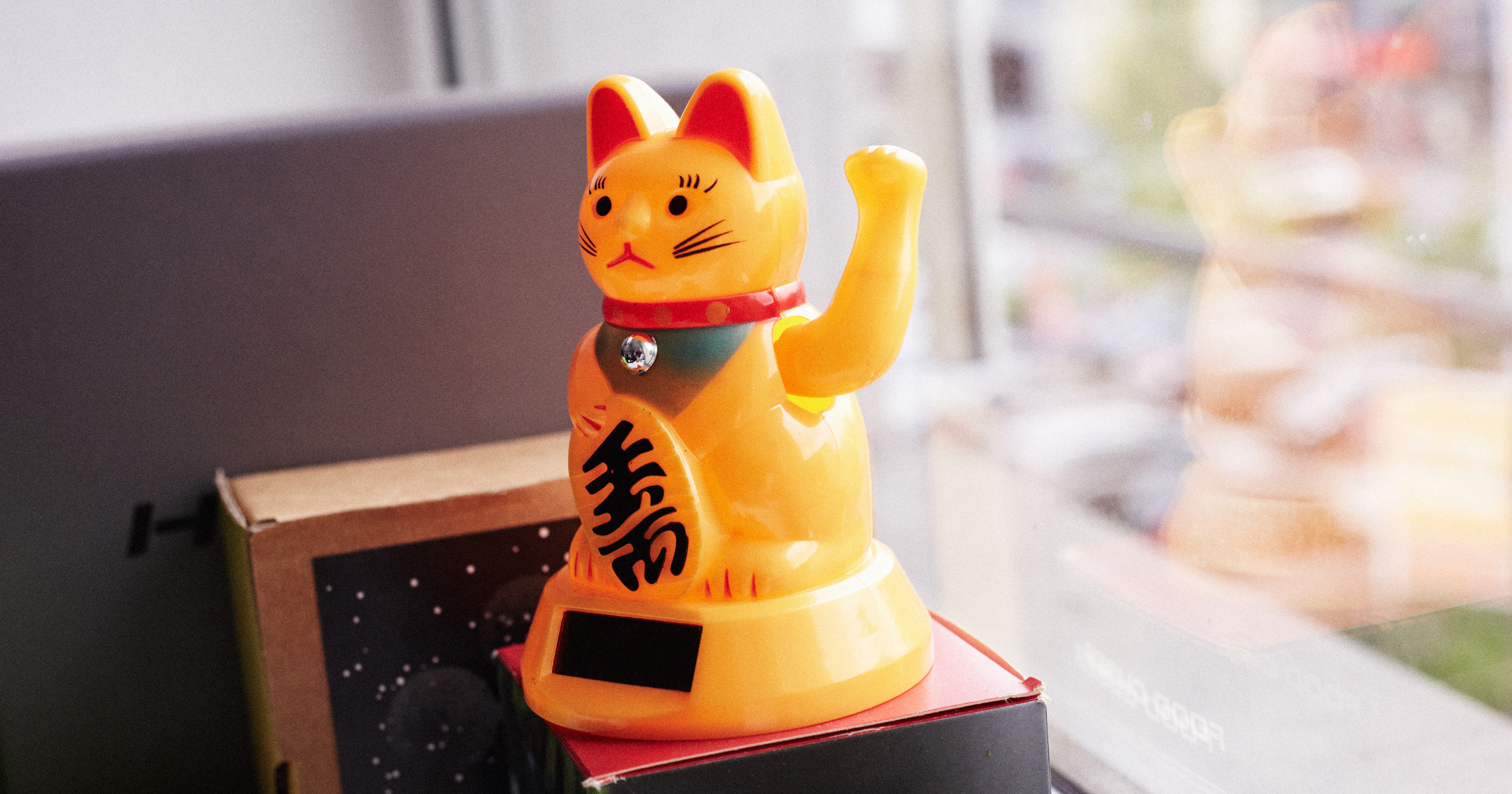 Das Bild zeigt eine orangene Winke-Katze aus Plastik, die ein einem Fenster steht.