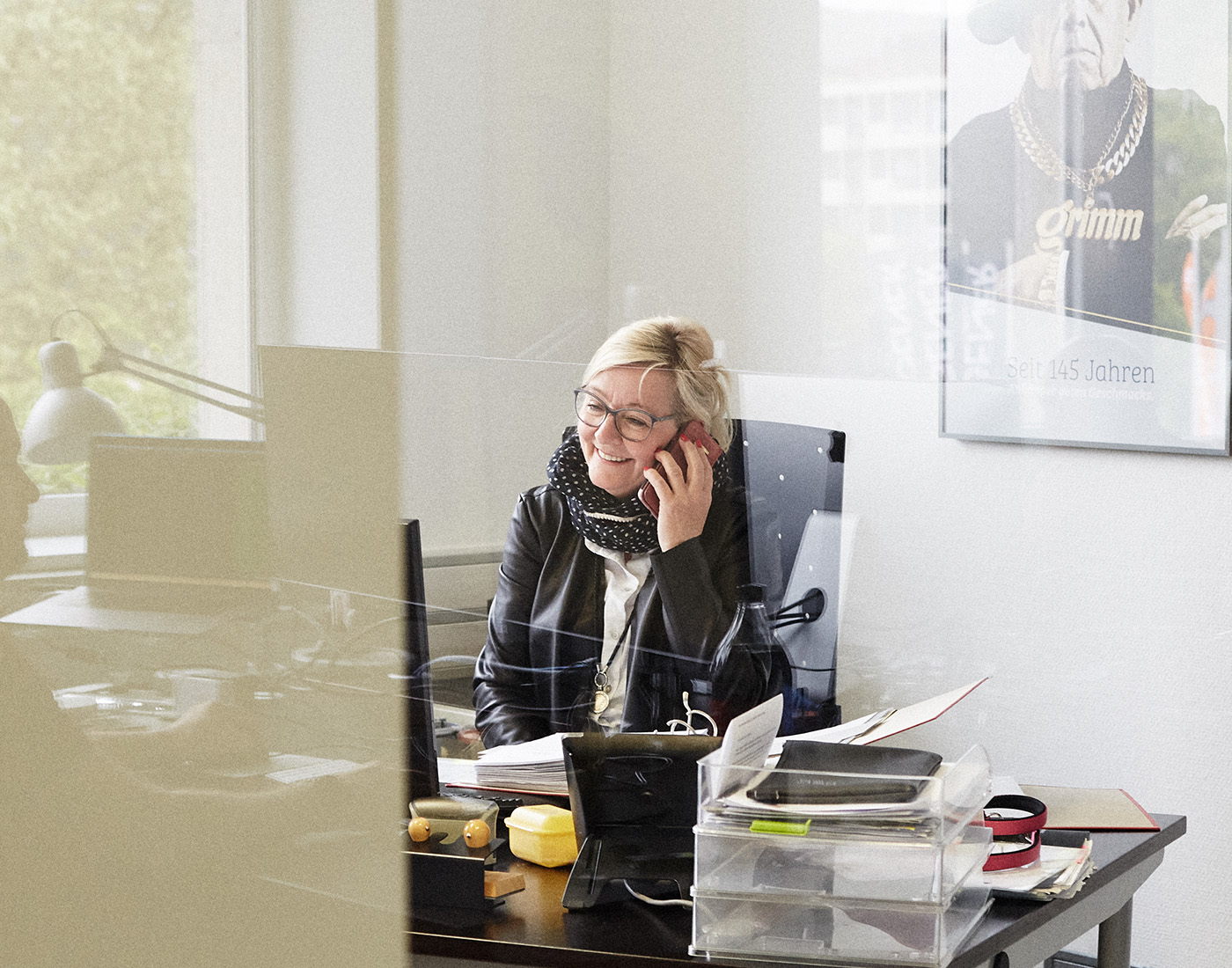 Dieses Bild zeigt eine FederhenSchneider Mitarbeiterin in den 40ern mit blonden Haaren und Brille. Sie sitzt am Schreibtisch, hält ein Smartphone an ihr Ohr und grinst gedankenverloren. Das Foto wurde durch eine Glaswand aufgenommen.