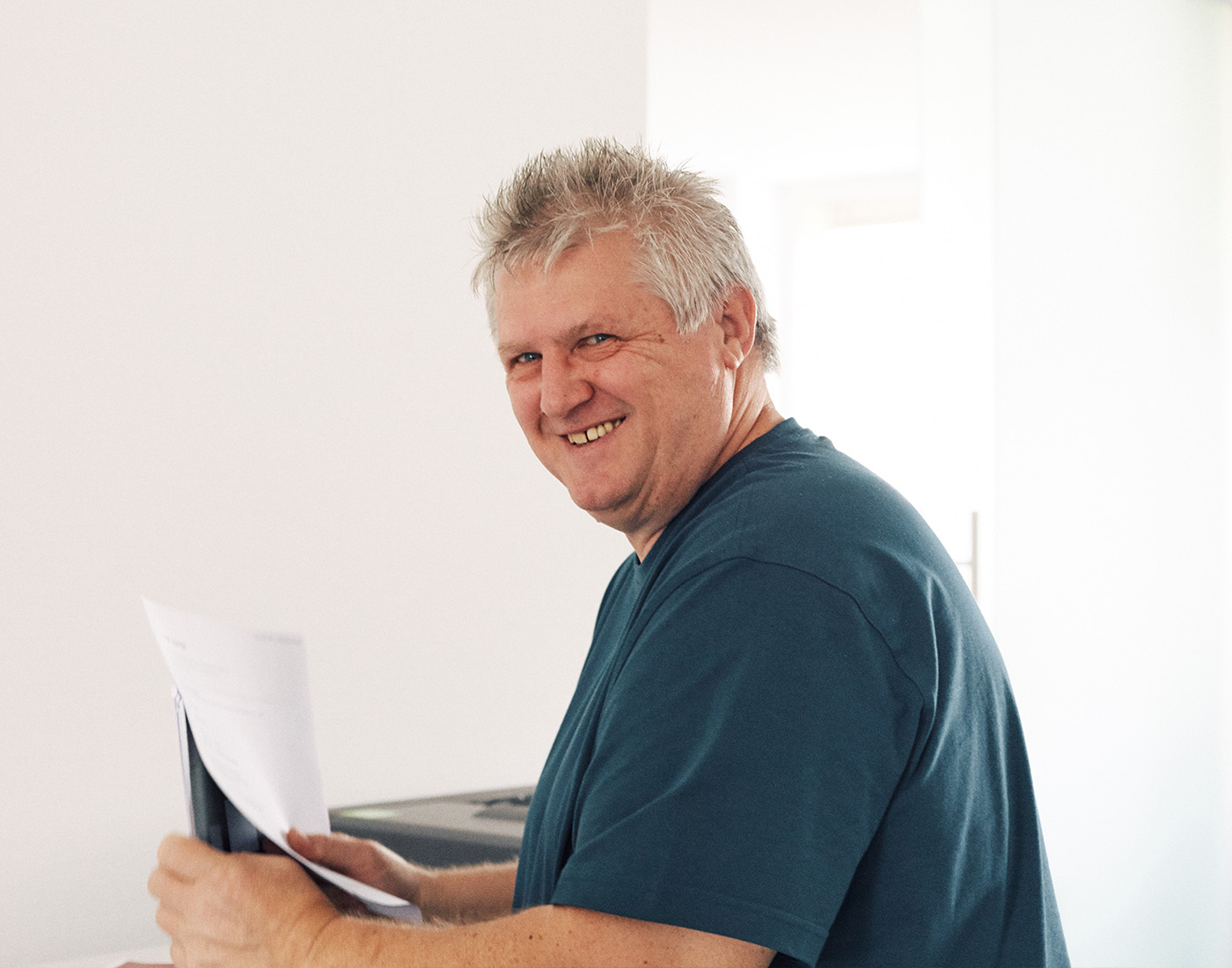 Dieses Bild zeigt einen FederhenSchneider Mitarbeiter in den 50ern mit grauen kurzen Haaren. Er hält ein Blatt Papier hoch während er seitlich in die Kamera grinst.