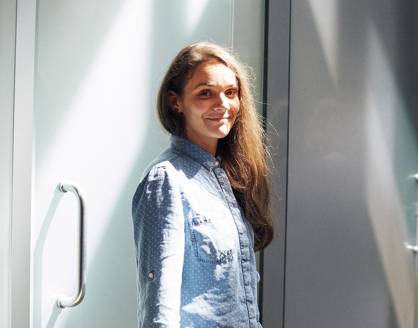 Dieses Bild zeigt eine FederhenSchneider Mitarbeiterin in den 20ern mit braunen langen Haaren, die seitlich über einer Schulter liegen. Über die andere Schulter lächelt sie in die Kamera während sie vor einer Milchglastür steht.
