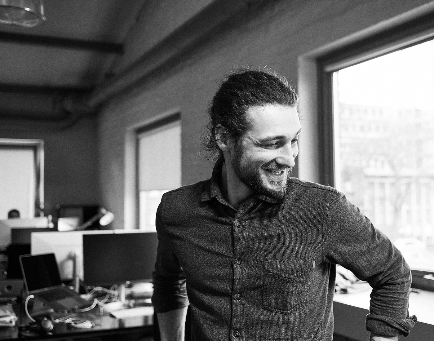 Dieses Bild zeigt einen FederhenSchneider Mitarbeiter in den 30ern mit Bart und langen dunklen Haaren zum Dutt. Er steht vor mehreren Schreibtischen, hat die Hände in die Hüpfte gestemmt, schaut leicht nach unten und lächelt an der Kamera vorbei.