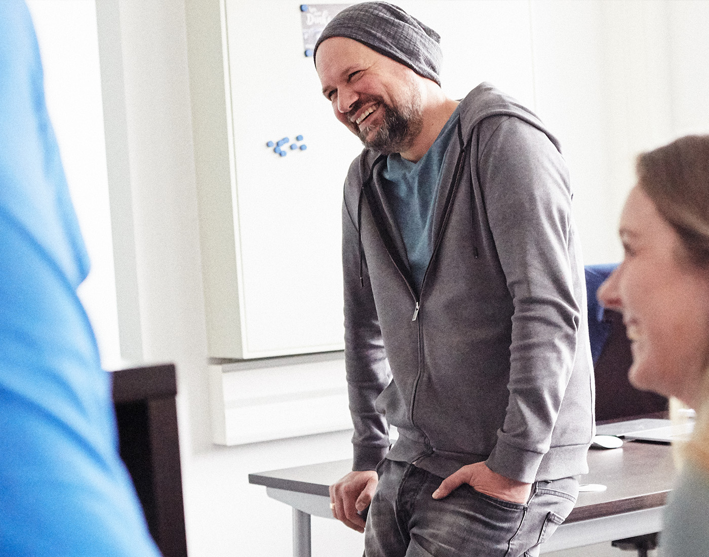 Dieses Bild zeigt einen der FederhenSchneider Geschäftsführer in den 40ern. Er lehnt an der Kante eines Schreibtisches, der hinter ihm steht und lächelt an der Kamera vorbei in den Raum. Im Anschnitt sieht man weitere FederhenSchneider Mitarbeiter, die sich mit ihm zu unterhalten scheinen.