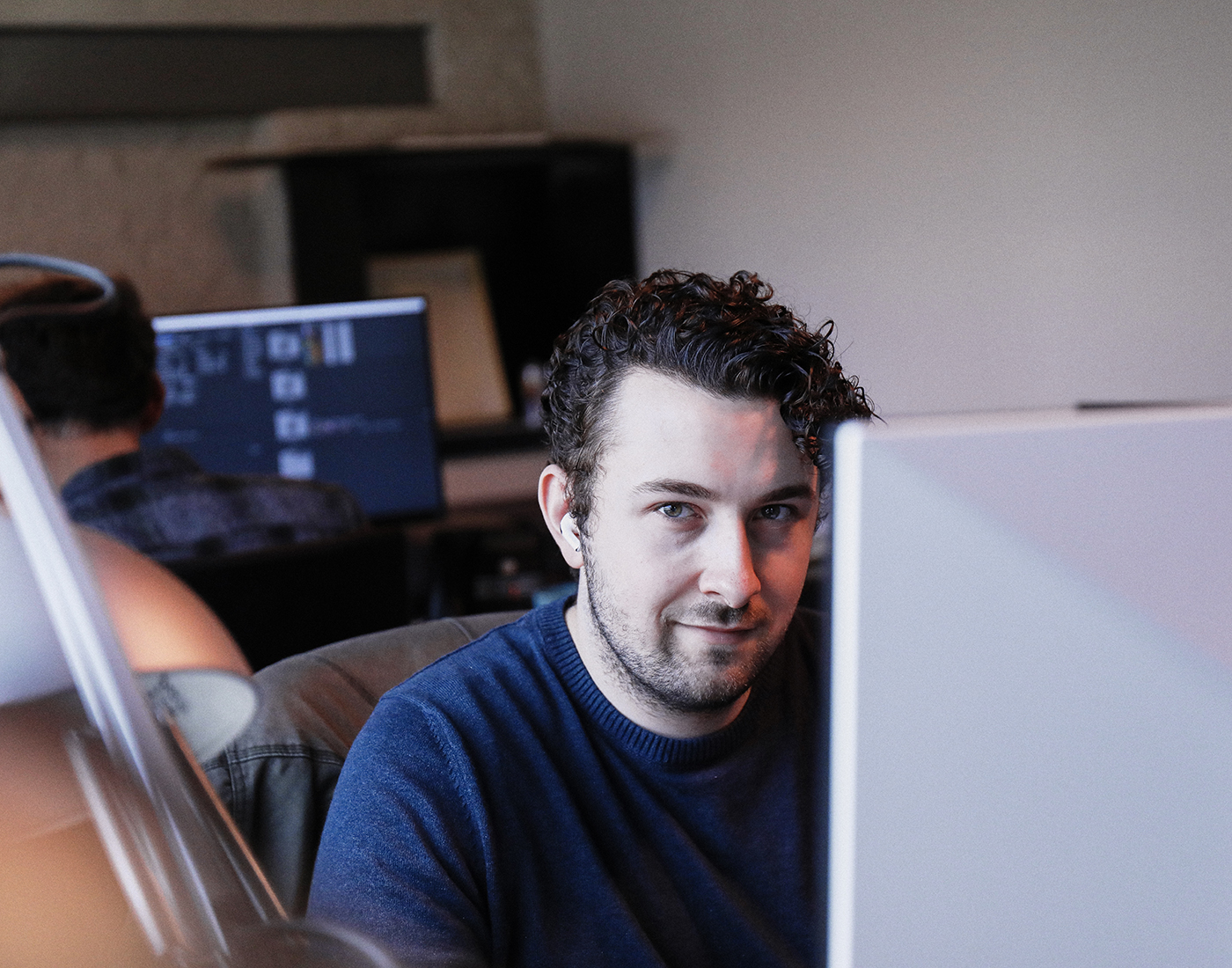 Dieses Bild zeigt einen FederhenSchneider Mitarbeiter Mitte 20 mit dunklen, gelockten Haaren und Bart, der an einem Monitor vorbei in die Kamera schaut und leicht lächelt. 