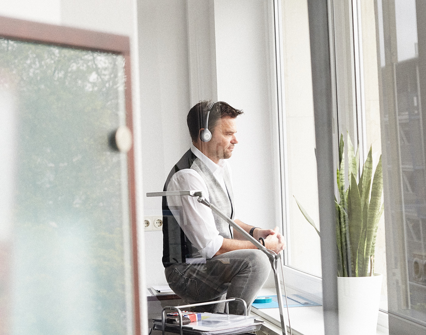 Dieses Bild zeigt einen der FederhenSchneider Geschäftsführer in den 50ern in seinem Büro. Er trägt Kopfhörer und schaut aus dem Fenster. Er steht seitlich zur Kamera und wirkt konzentriert. Das Foto wurde durch eine Glasscheibe aufgenommen.