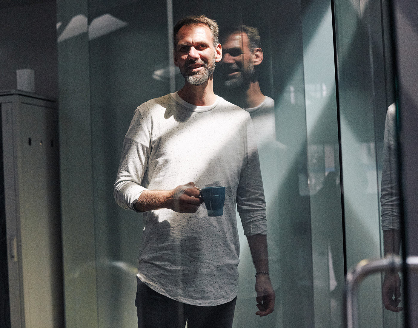 Dieses Bild zeigt einen FederhenSchneider Mitarbeiter in den 40ern mit Kaffeetasse, der  in die Kamera lächelt. Er steht vor einer Glaswand, hinter ihm eine Glaswand. Das Foto ist durch eine Glaswand aufgenommen, sodass seine Silhouette sich darin spiegelt.