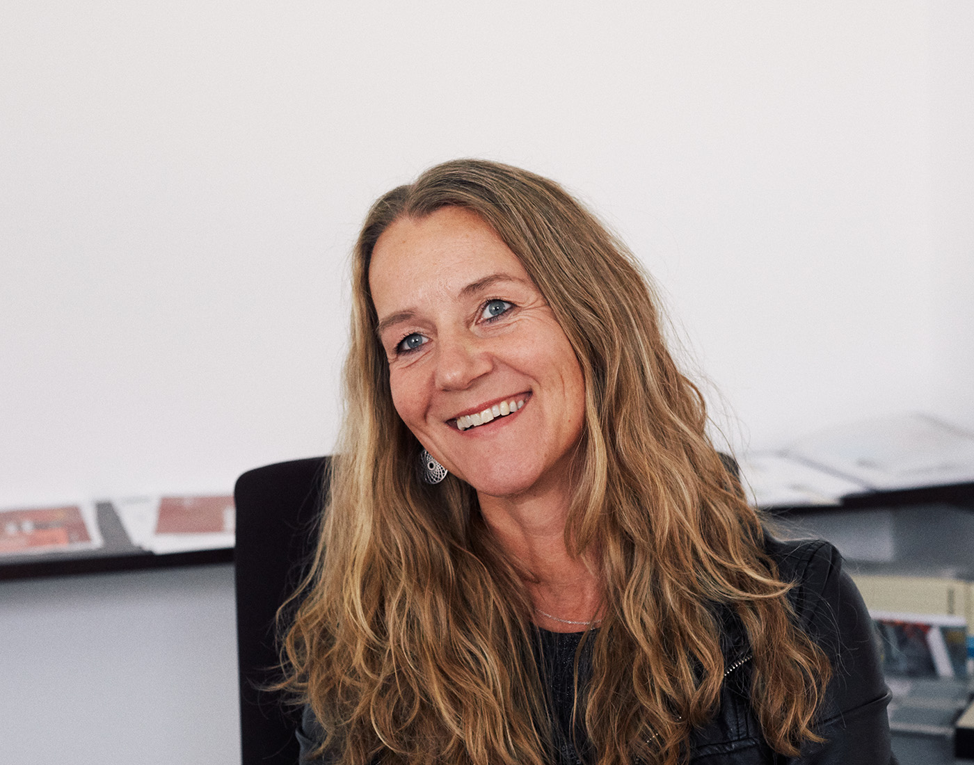 Dieses Bild zeigt eine FederhenSchneider Mitarbeiterin in den 40ern mit blonden, lockigen Haaren, die auf einem Schreibtischstuhl sitzt und mit leicht schräg gelegtem Kopf an der Kamera vorbei lächelt.