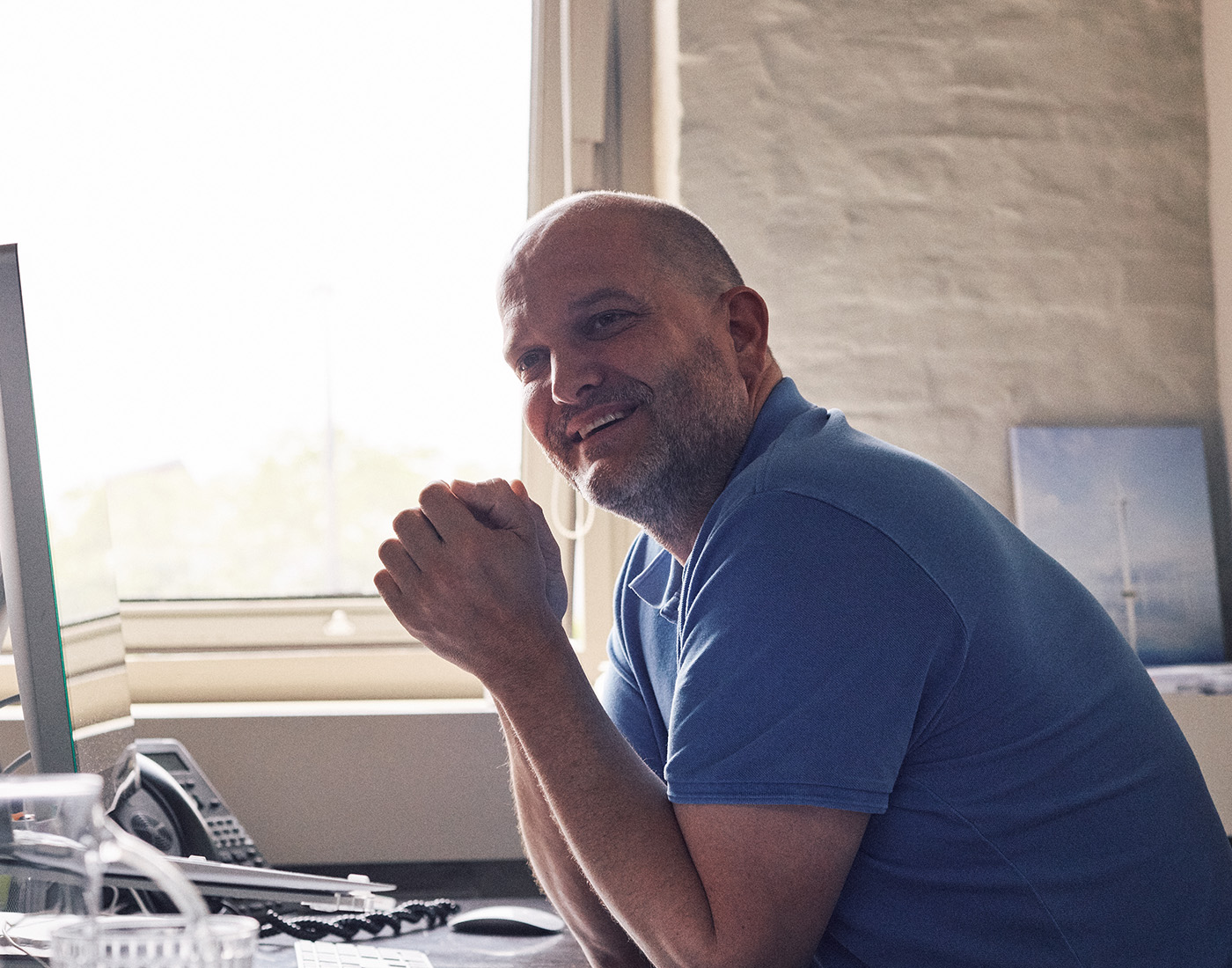 Dieses Bild zeigt einen FederhenSchneider Mitarbeiter in den 40ern mit Glatze und grauem Bart an seinem Schreibtisch, von der Seite fotografiert. Er hat beide Ellbogen auf den Tisch gestützt und schaut amüsiert an der Kamera vorbei.