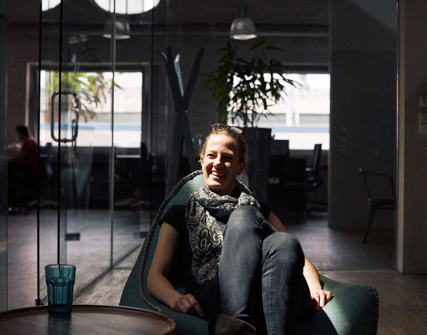 Dieses Bild zeigt eine FederhenSchneider Mitarbeiterin in den 40ern mit Brille auf dem Kopf. Sie sitzt auf einem türkisene Sitzsack mit übereinandergeschlagenen Beinen. Ein Lichtkegel fällt über ihr Gesicht. Sie grinst an der Kamera vorbei.