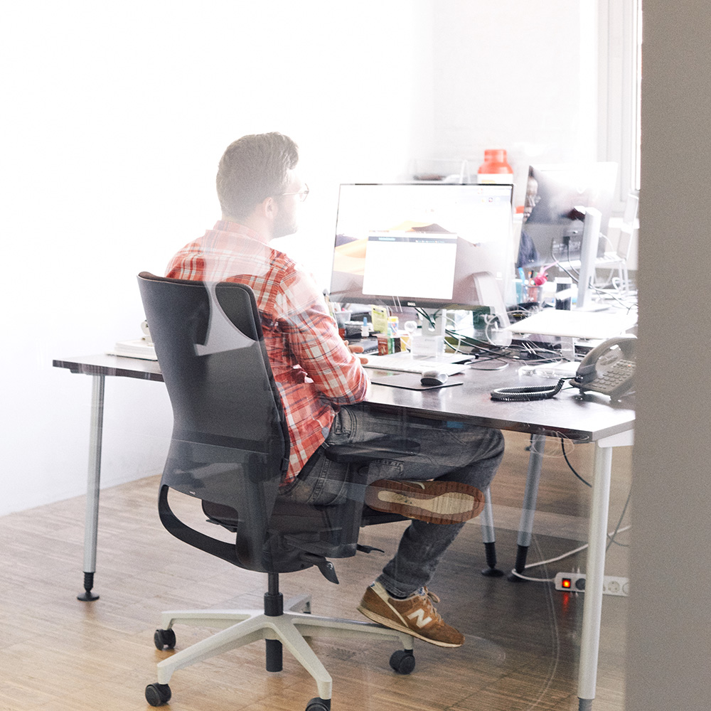 Dieses Bild zeigt einen FederhenSchneider Mitarbeiter am Schreibtisch, der auf seinen Monitor schaut. Das Bild ist von hinten durch eine Glaswand aufgenommen.