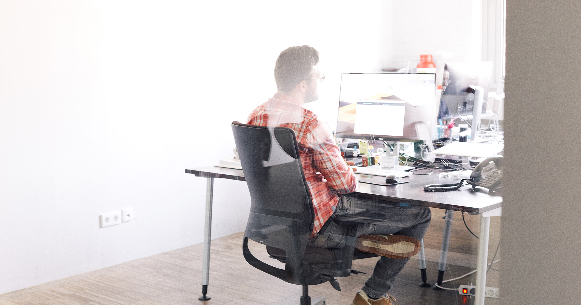 Dieses Bild zeigt einen FederhenSchneider Mitarbeiter am Schreibtisch, der auf seinen Monitor schaut. Das Bild ist von hinten durch eine Glaswand aufgenommen.