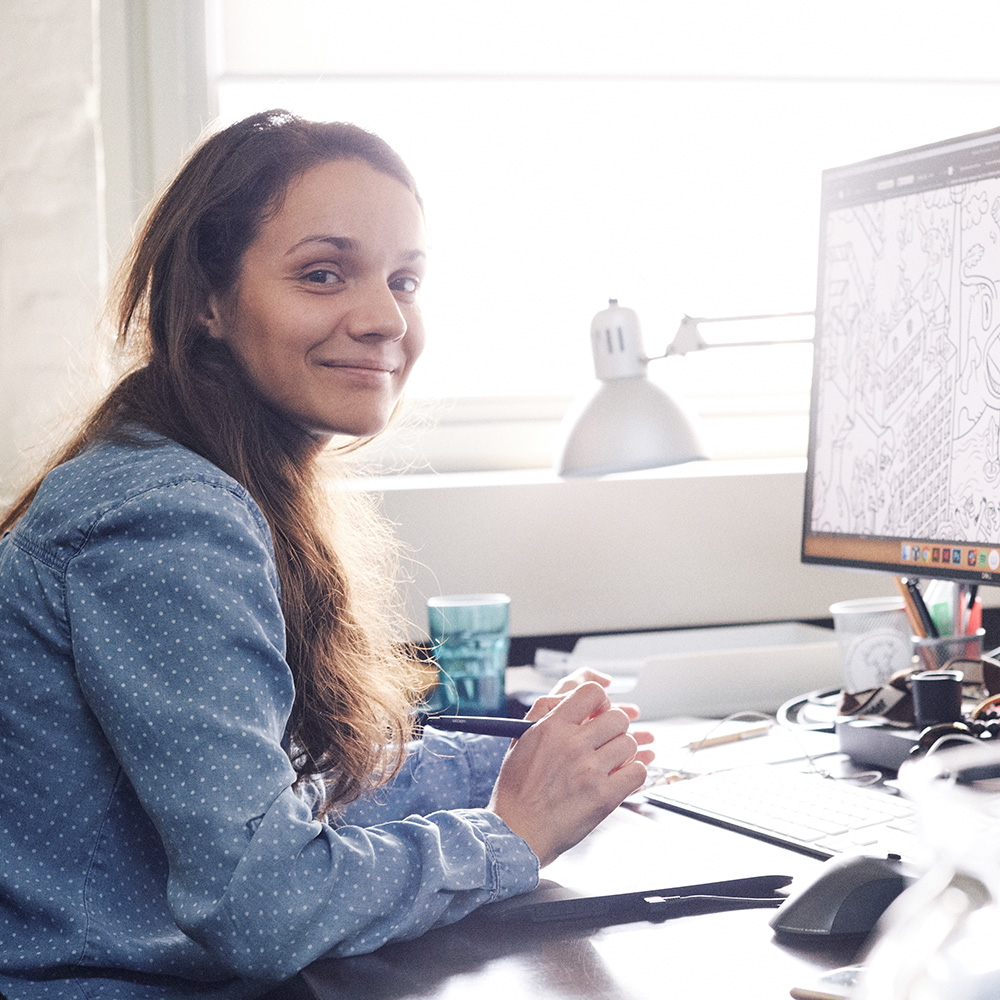 Dieses Bild zeigt eine FederhenSchneider Mitarbeiterin am Schreibtisch, die seitlich in die Kamera schaut. Auf ihrem Monitor ist eine Comic-Illustration zu sehen. 