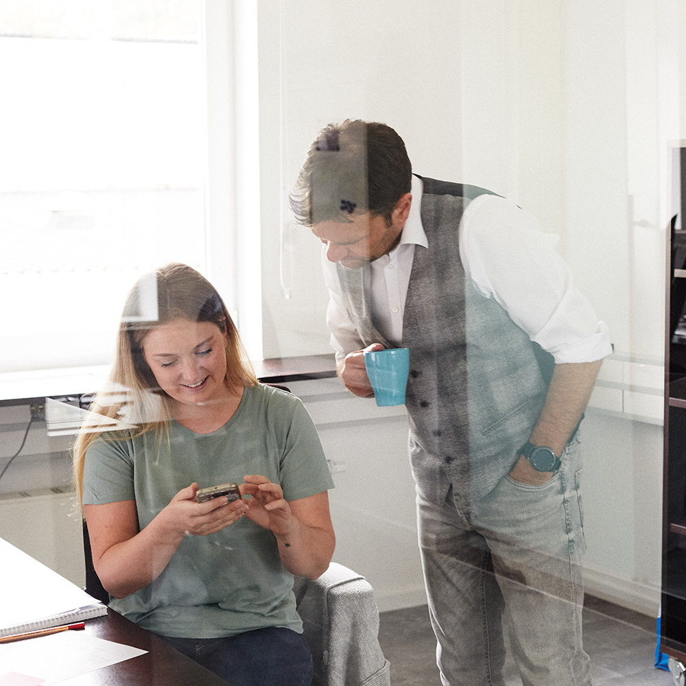 Dieses Bild zeigt eine FederhenSchneider Mitarbeiterin, die einem der FederhenSchneider Geschäftsführer etwas auf dem Smartphone zeigt. Das Foto ist durch eine Glaswand aufgenommen.