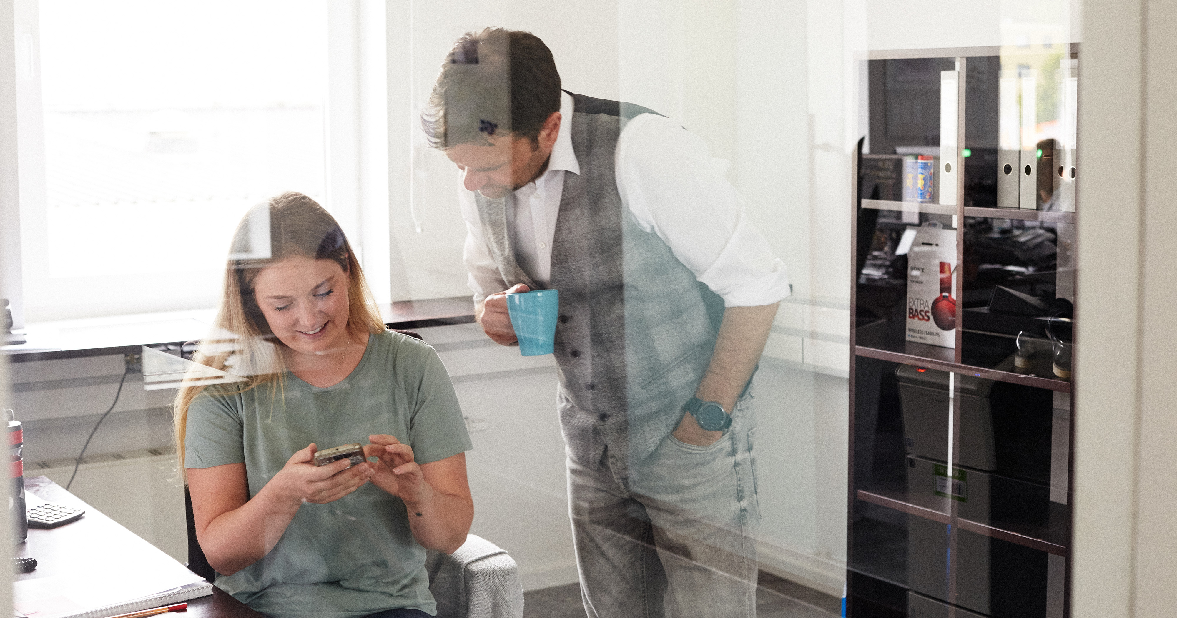Dieses Bild zeigt eine FederhenSchneider Mitarbeiterin, die einem der FederhenSchneider Geschäftsführer etwas auf dem Smartphone zeigt. Das Foto ist durch eine Glaswand aufgenommen.