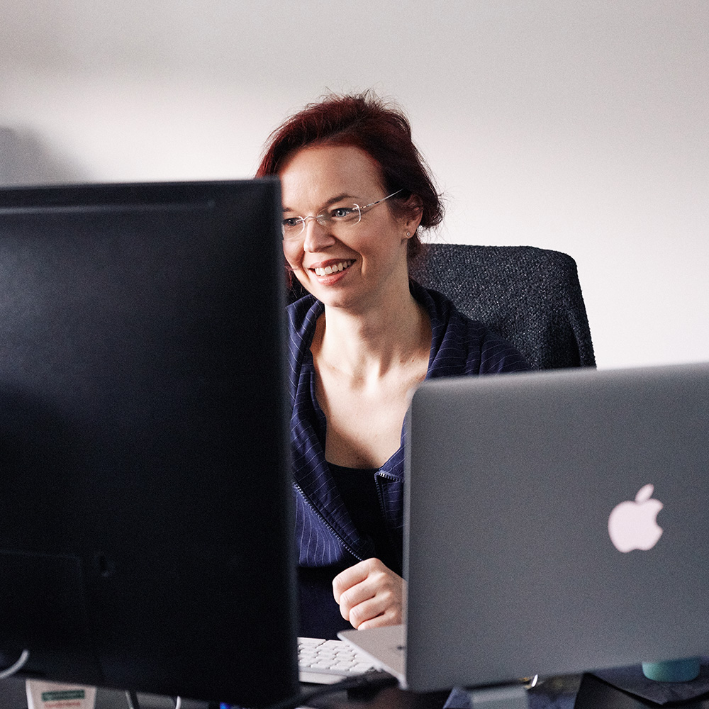 Dieses Bild zeigt eine FederhenSchneider Mitarbeiterin, die zwischen zwei Monitoren hindurch bei der Arbeit fotografiert wurde.