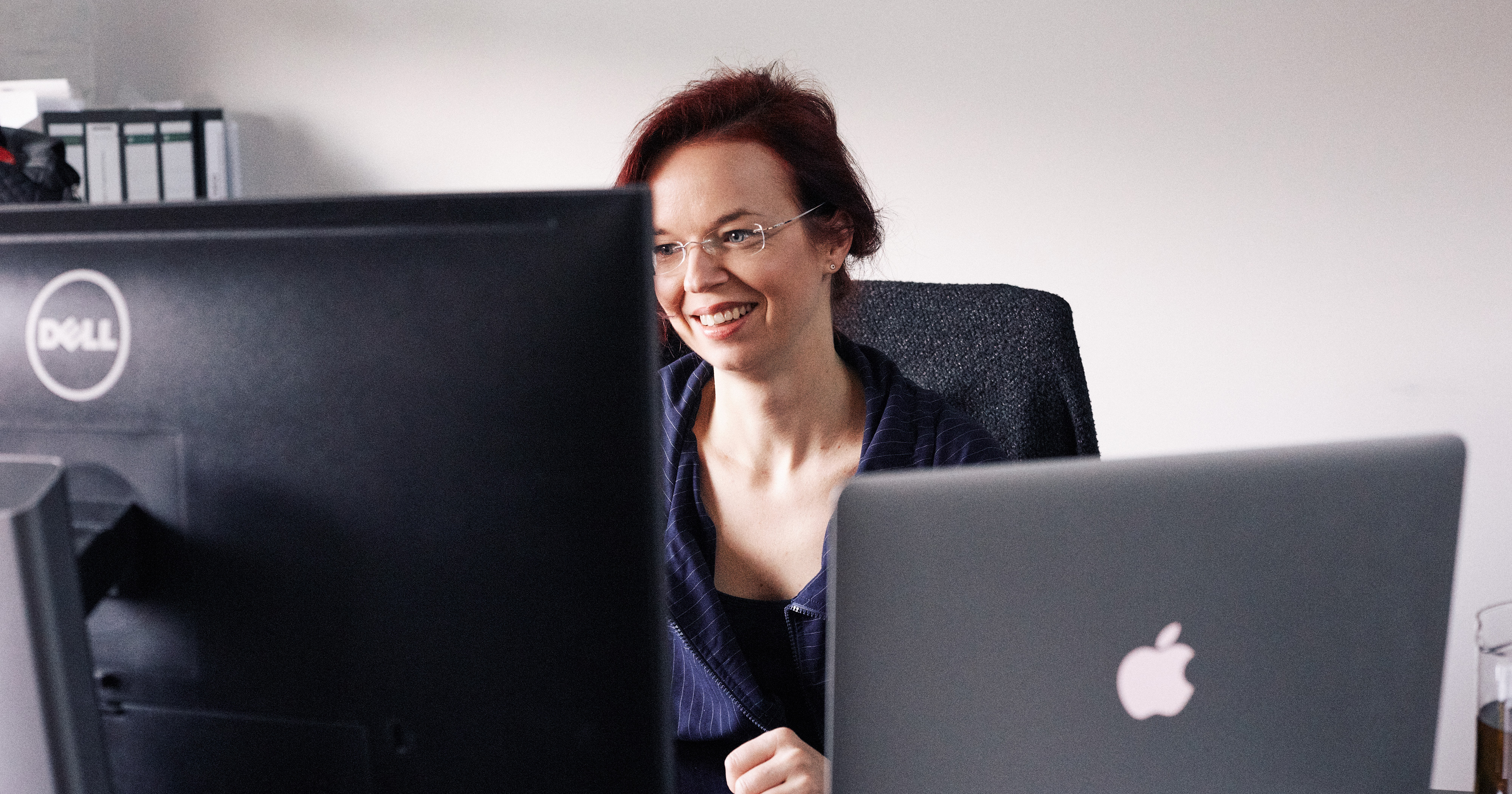 Dieses Bild zeigt eine FederhenSchneider Mitarbeiterin, die zwischen zwei Monitoren hindurch bei der Arbeit fotografiert wurde.