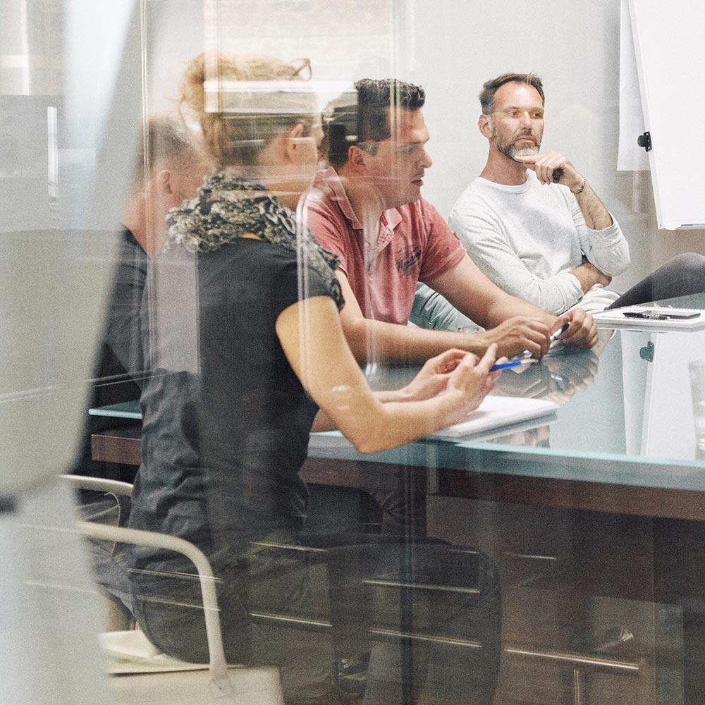 Ein Team von Produktionsexperten bei einer Besprechung, durch eine Glasscheibe fotografiert. Die Gruppe sitzt um einen Konferenztisch, notiert und diskutiert aufmerksam.