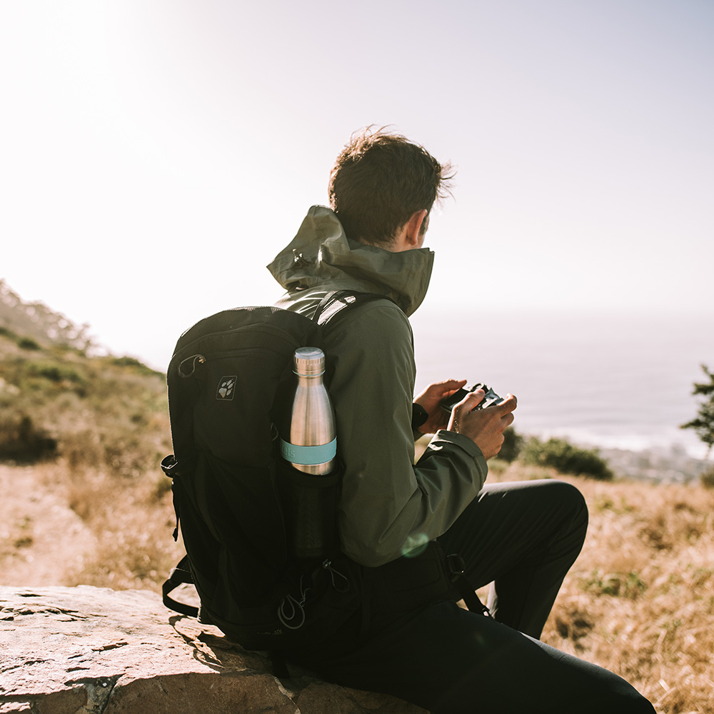 Das Foto zeigt einen Mann von hinten, der in der Natur auf einem Stein sitzt von hinten. An seinem Rucksack sieht man seitlich eine Bodddelsflasche.