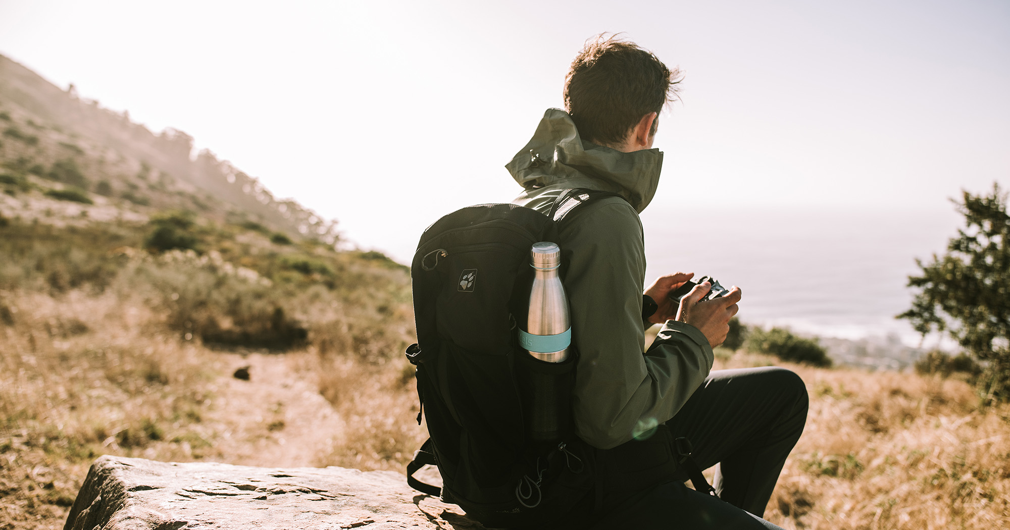 Das Foto zeigt einen Mann von hinten, der in der Natur auf einem Stein sitzt von hinten. An seinem Rucksack sieht man seitlich eine Bodddelsflasche.