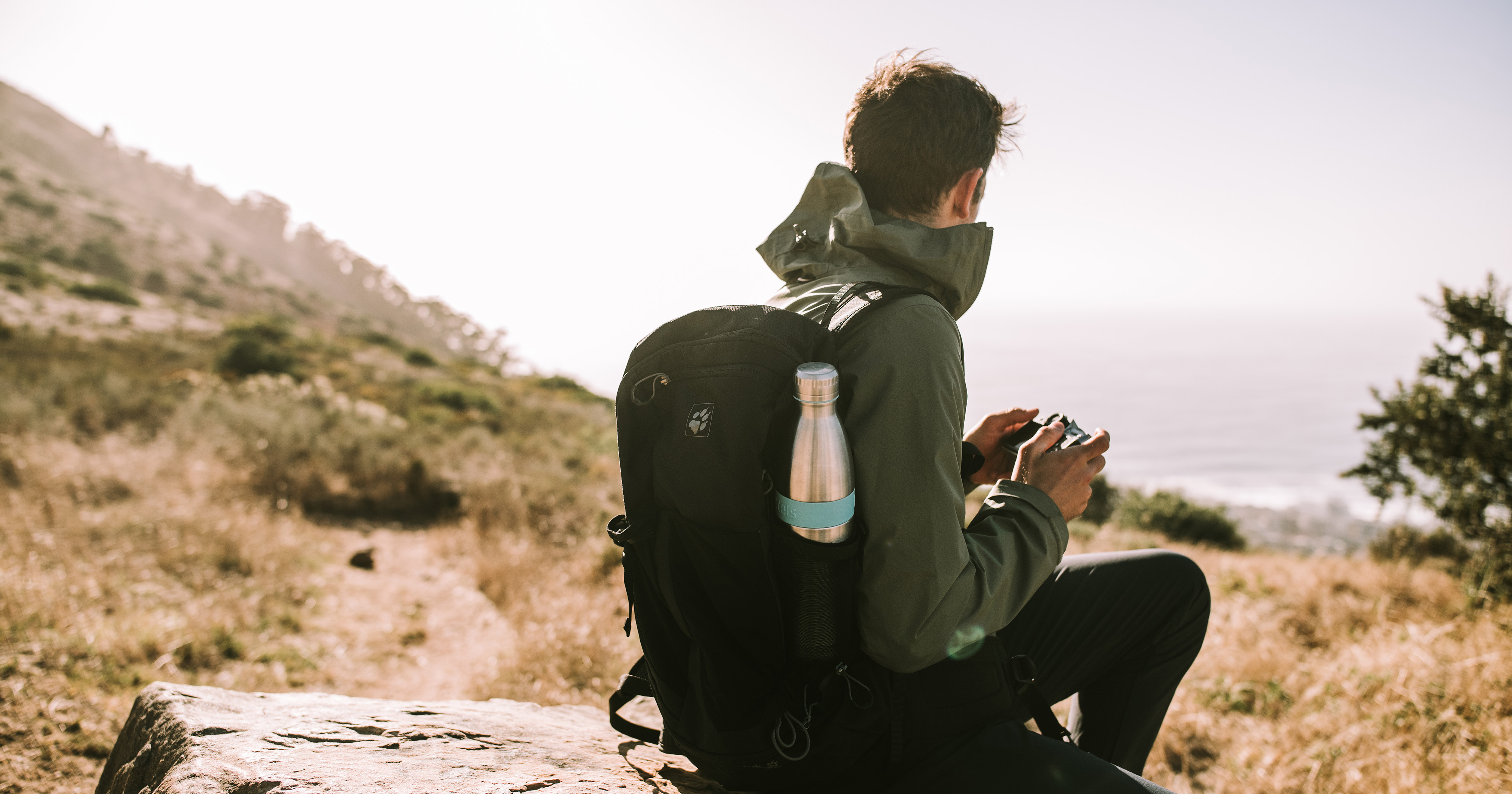 Das Foto zeigt einen Mann von hinten, der in der Natur auf einem Stein sitzt von hinten. An seinem Rucksack sieht man seitlich eine Bodddelsflasche.