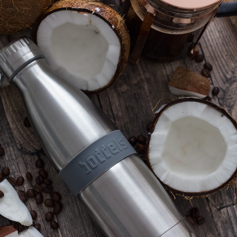 Das Foto zeigt eine Boddels TWEE Trinkflasche in grau nebst Kokosnüssen auf einer Tischplatte aus dunklem Holz.