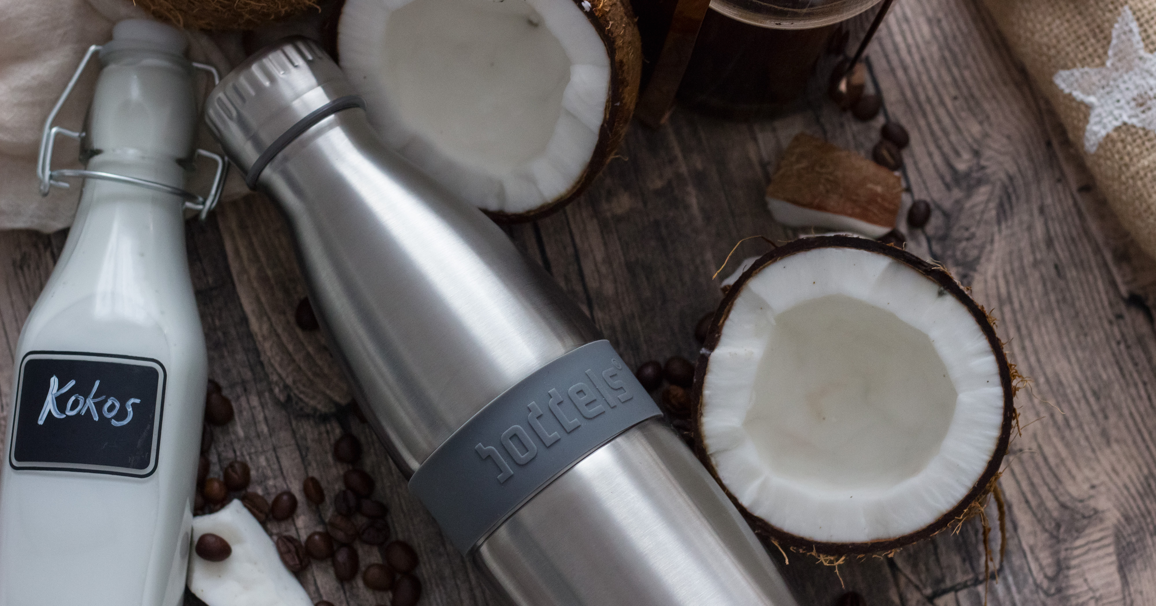 Das Foto zeigt eine Boddels TWEE Trinkflasche in grau nebst Kokosnüssen auf einer Tischplatte aus dunklem Holz.