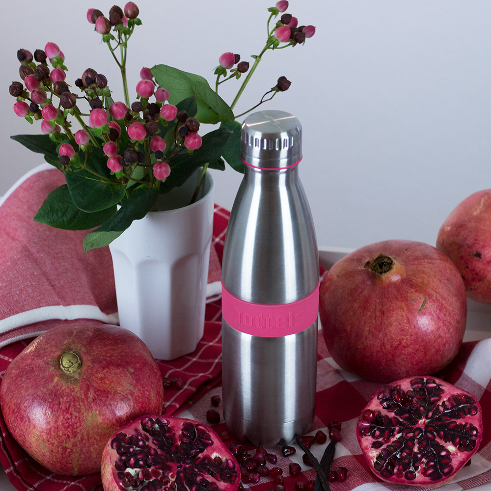 Das Foto zeigt eine Boddels Trinkflasche in pink nebst Granatäpfeln und pinken Blumen in einer Vase.