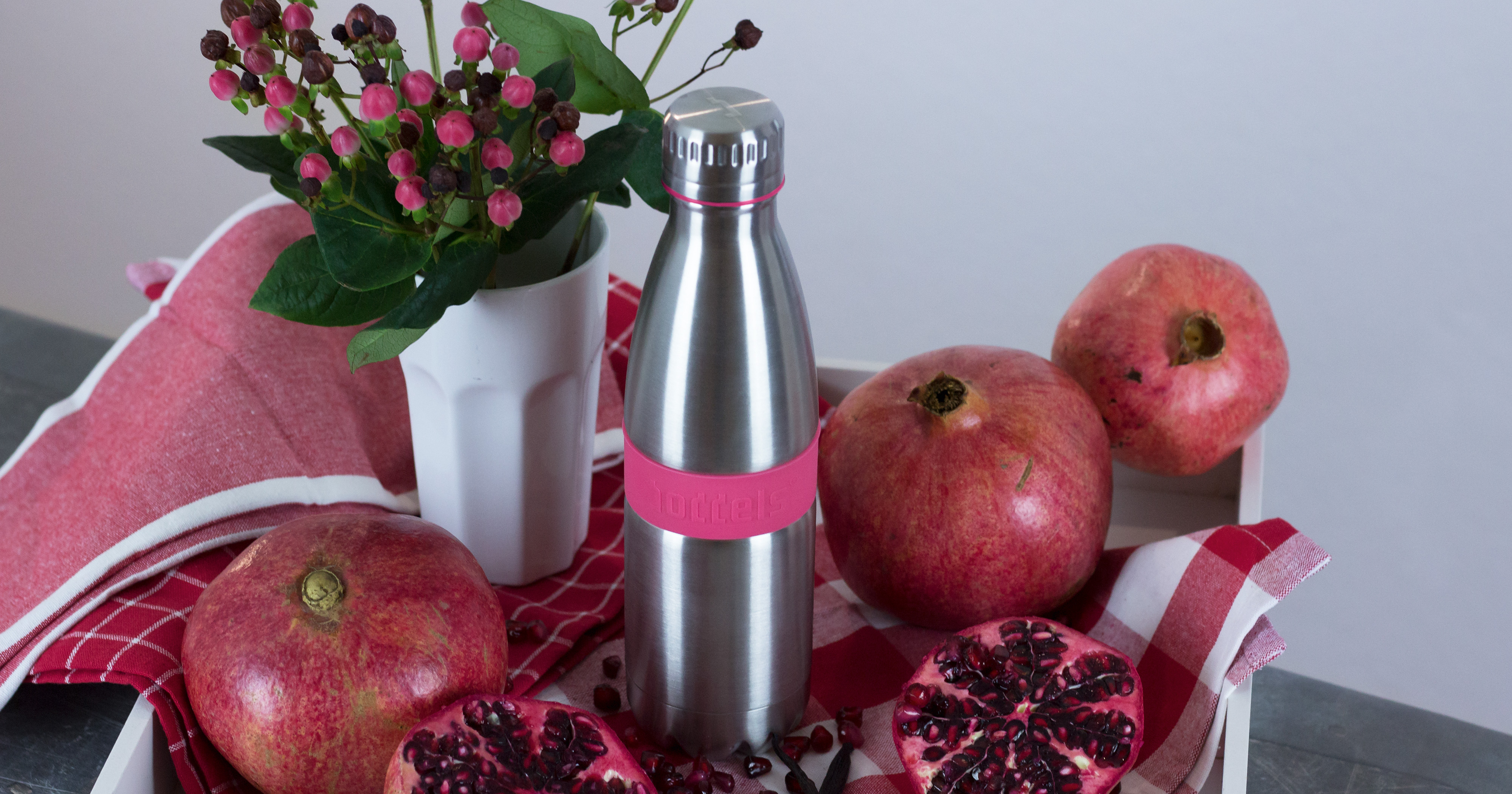 Das Foto zeigt eine Boddels Trinkflasche in pink nebst Granatäpfeln und pinken Blumen in einer Vase.
