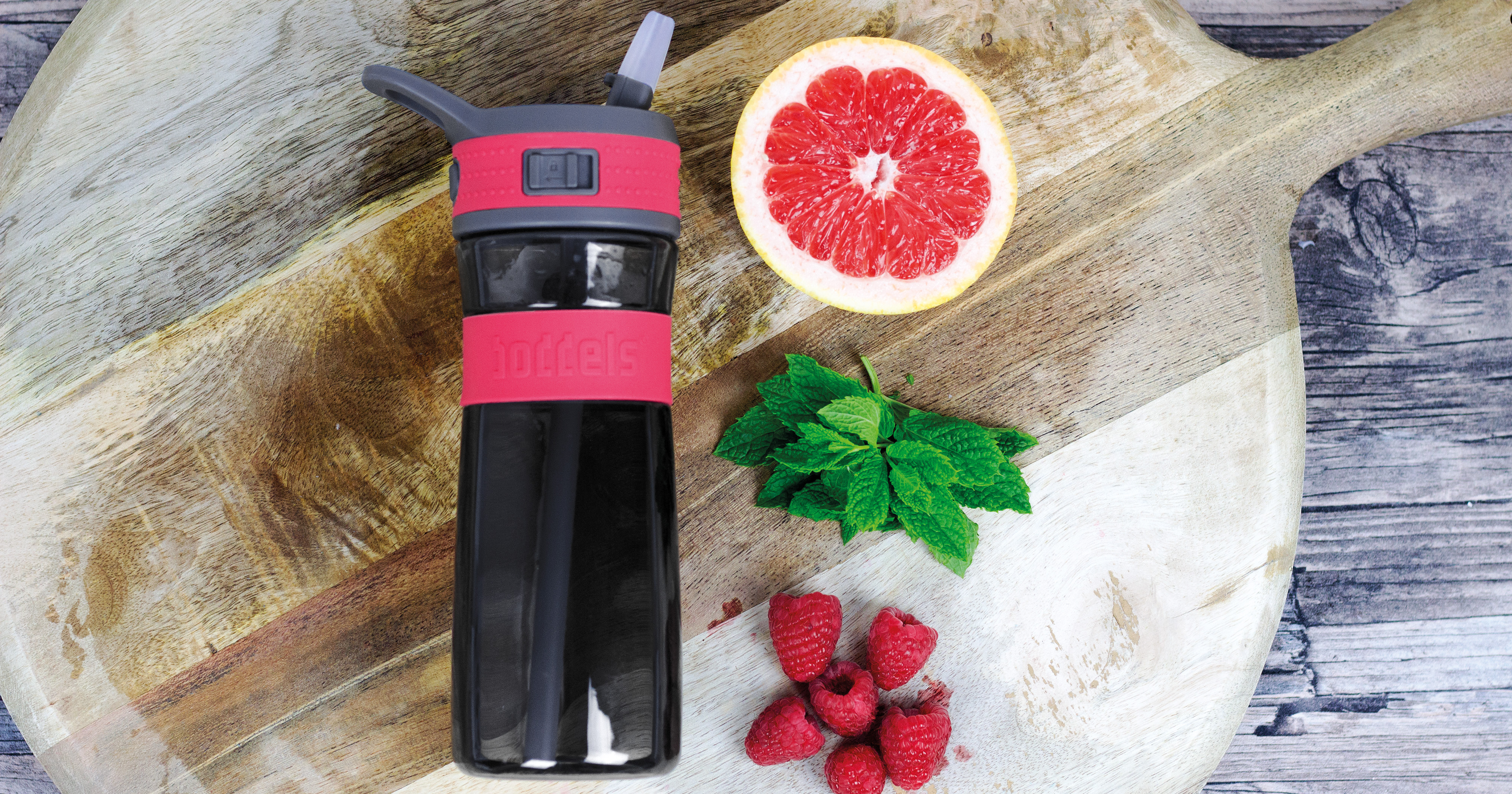 Das Foto zeigt eine Boddels Trinkflasche in pink nebst Grapefruit, Minzblättern und Himbeeren auf einem Schneidebrett aus Holz liegend.