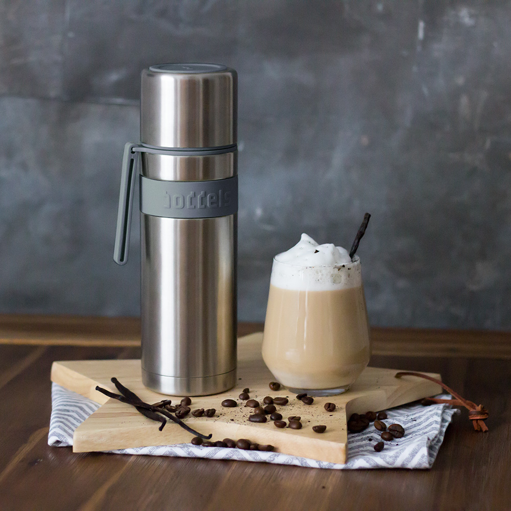Das Foto zeigt eine Boddels Trinkflasche in grau nebst Glas mit Sahnekaffe und Strohhalm und diversen Gewürzen auf einem Holzbrett in Sternform.