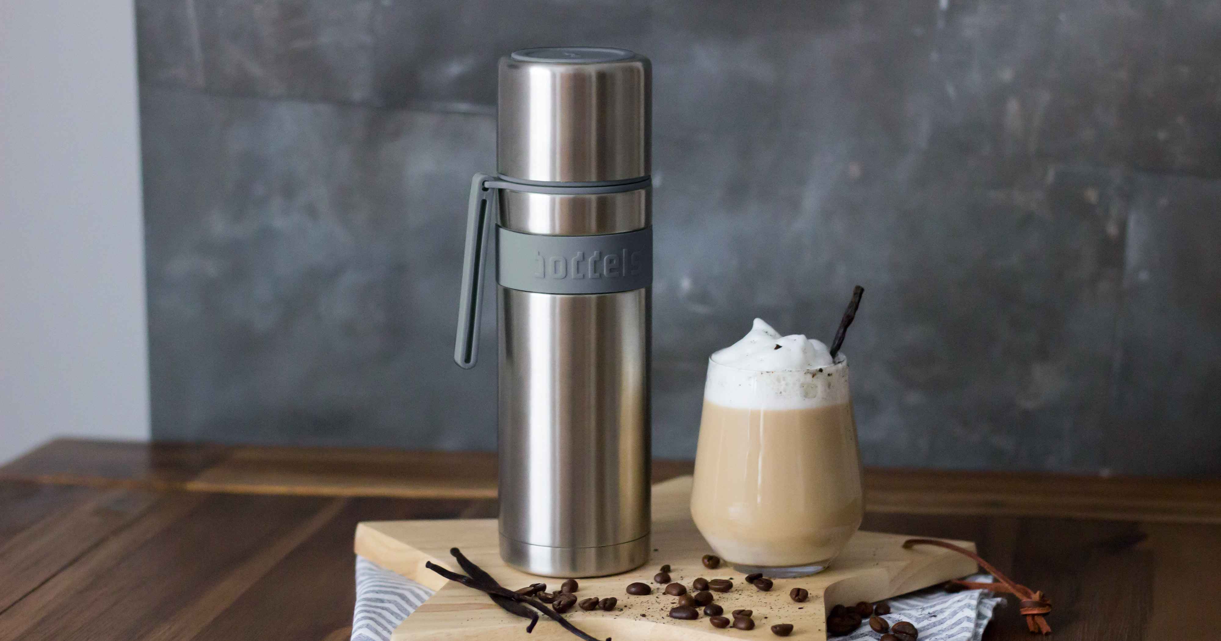 Das Foto zeigt eine Boddels Trinkflasche in grau nebst Glas mit Sahnekaffe und Strohhalm und diversen Gewürzen auf einem Holzbrett in Sternform.