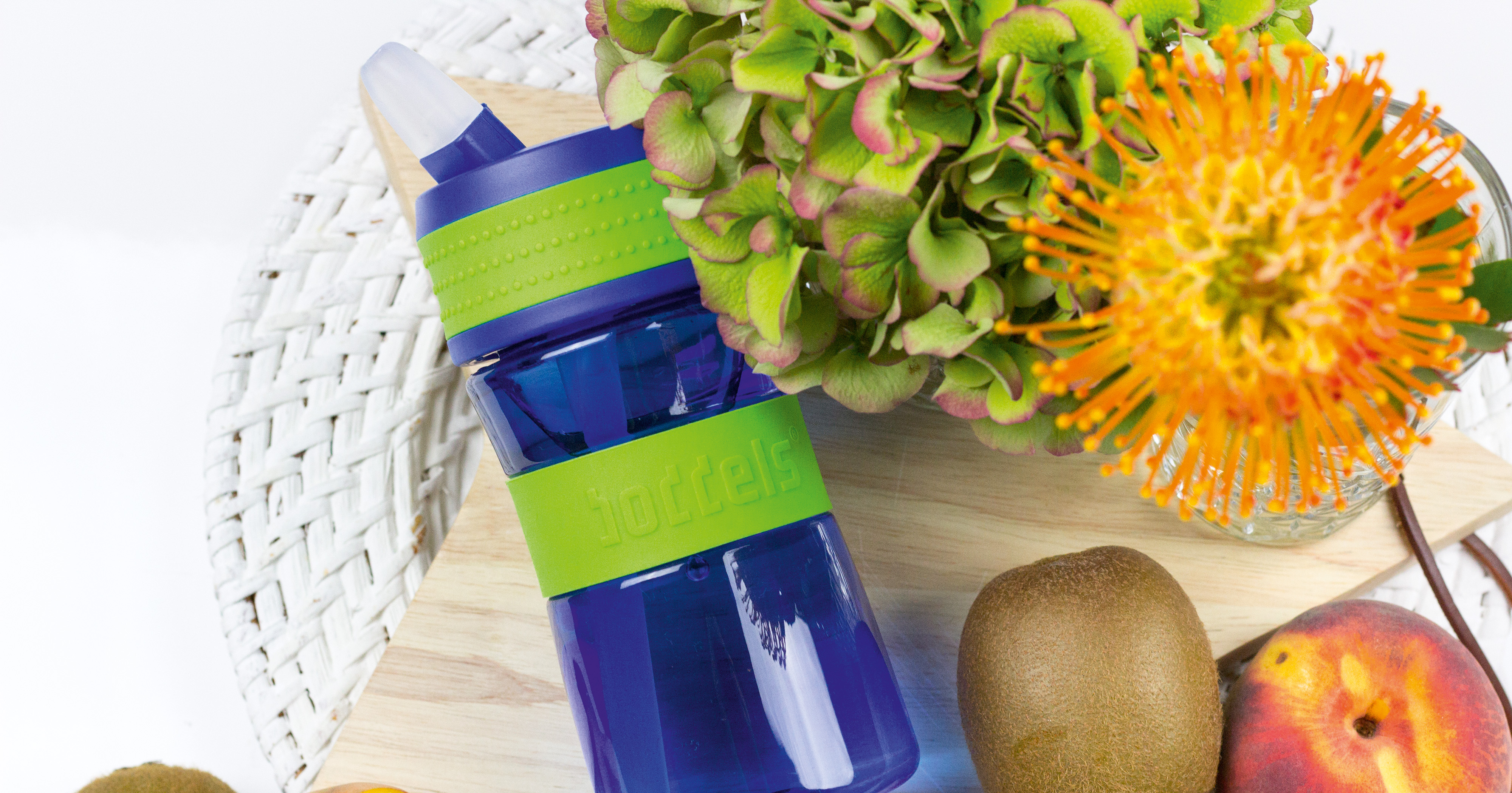 Das Foto zeigt eine Boddels Trinkflasche in grün nebst diversem Obst auf einer weißen Rattan-Oberfläche liegend.