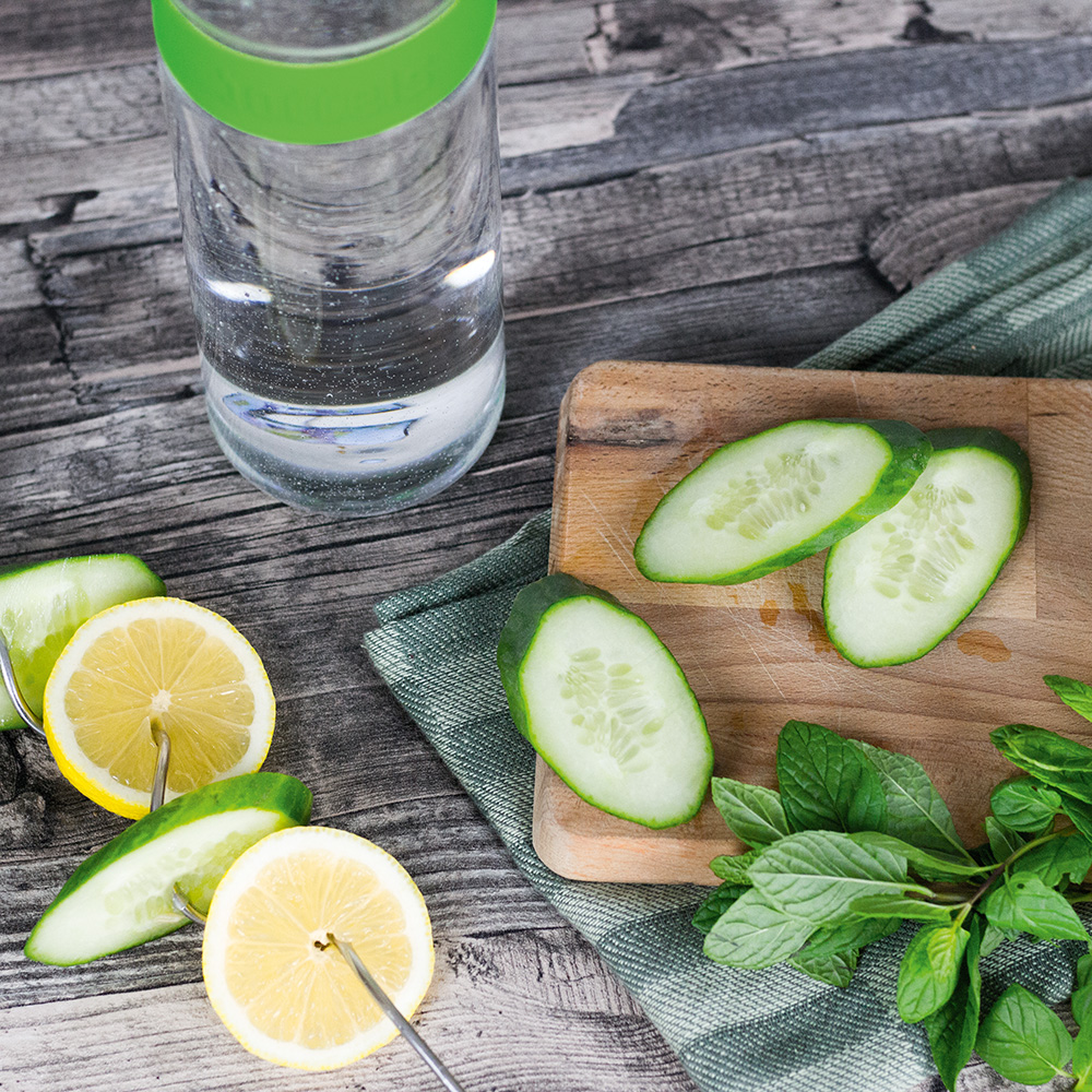 Das Foto zeigt eine Boddels Glaskaraffe in grün mit danebenliegendem Fruchtspieß gespickt mit Scheiben von Zitrone und Gurke im Wechsel.