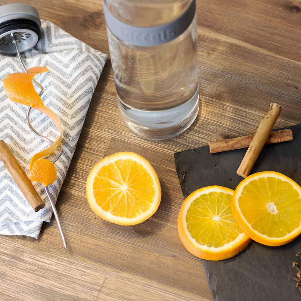 Das Foto zeigt eine Boddels Glaskaraffe in grau mit danebenliegendem Fruchtspieß um den Orangenzeste gewickelt ist, daneben Zimtstangen.