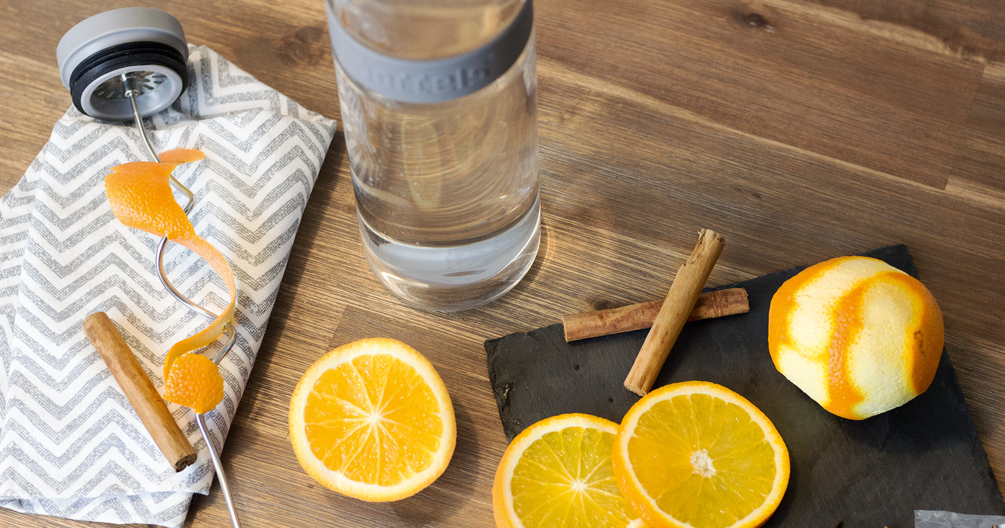 Das Foto zeigt eine Boddels Glaskaraffe in grau mit danebenliegendem Fruchtspieß um den Orangenzeste gewickelt ist, daneben Zimtstangen.