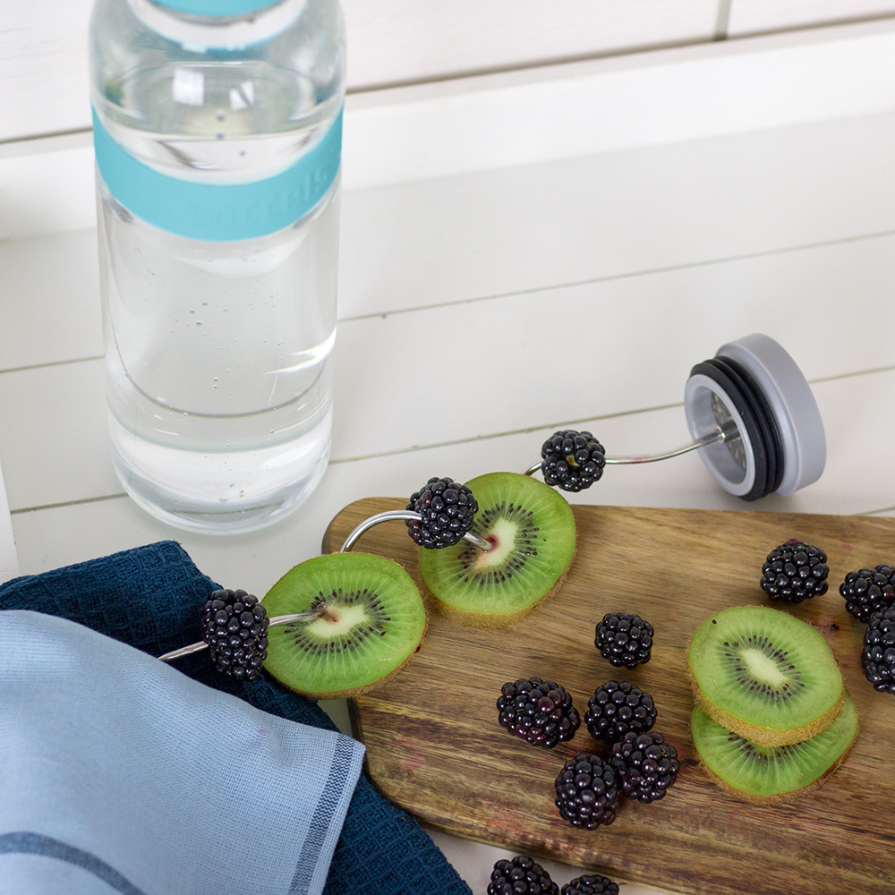 Das Foto zeigt eine Boddels Glaskaraffe in babyblau mit danebenliegendem Fruchtspieß gespickt mit Scheiben von Kiwi und Heidelbeeren im Wechsel.