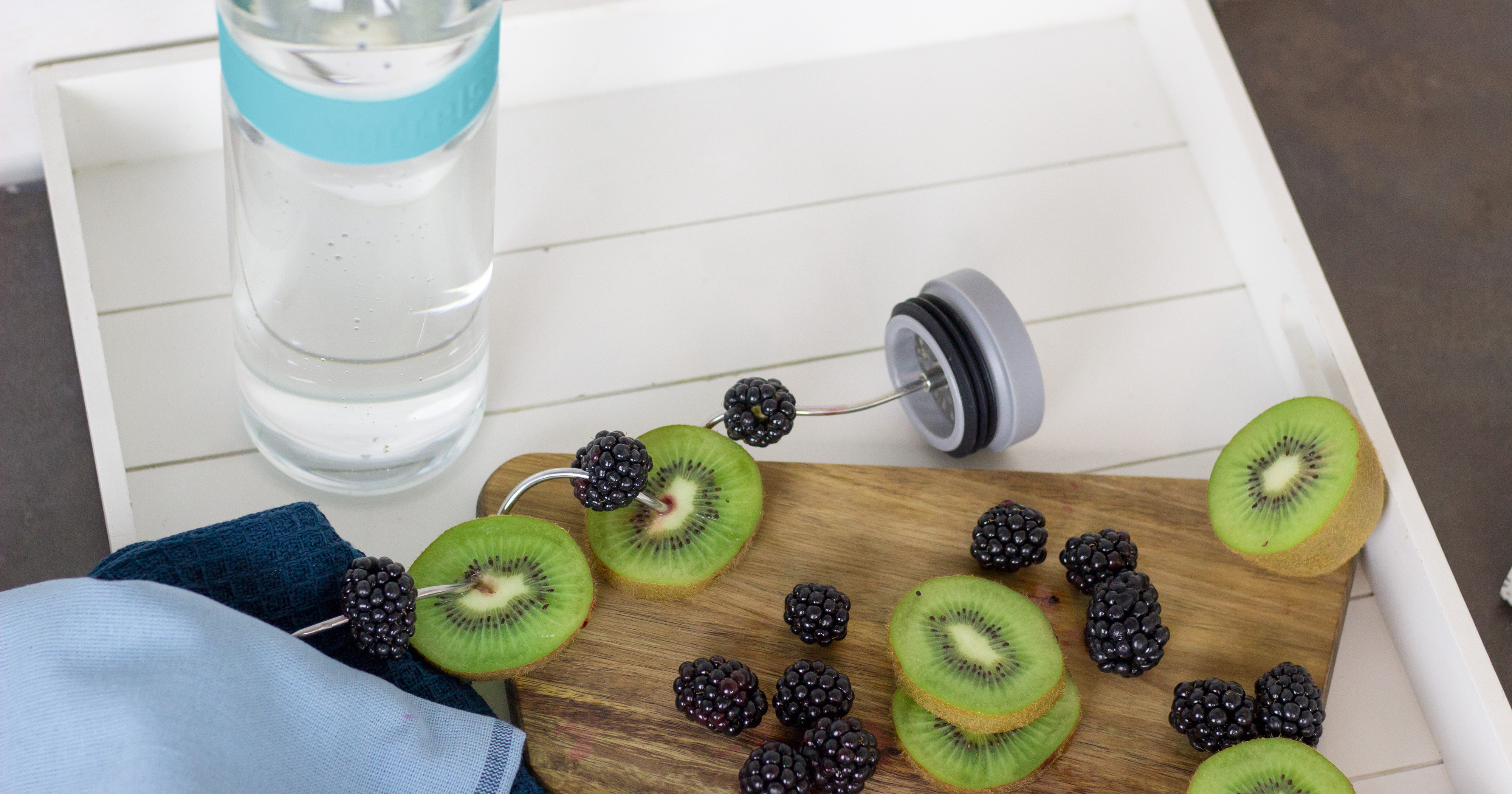 Das Foto zeigt eine Boddels Glaskaraffe in babyblau mit danebenliegendem Fruchtspieß gespickt mit Scheiben von Kiwi und Heidelbeeren im Wechsel.