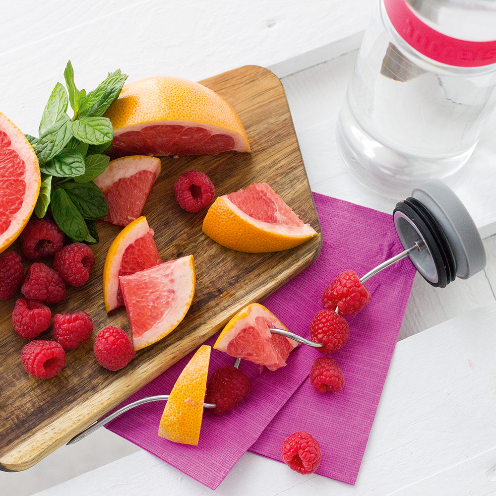 Das Foto zeigt eine Boddels Glaskaraffe in pink mit danebenliegendem Fruchtspieß gespickt mit Grapefruitstückchen und Himbeeren im Wechsel.
