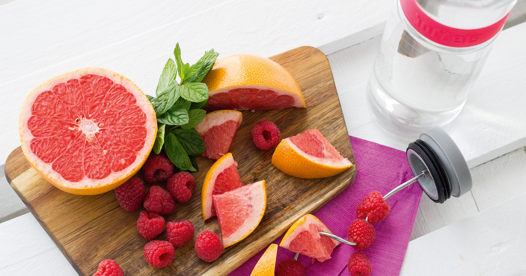 Das Foto zeigt eine Boddels Glaskaraffe in pink mit danebenliegendem Fruchtspieß gespickt mit Grapefruitstückchen und Himbeeren im Wechsel.