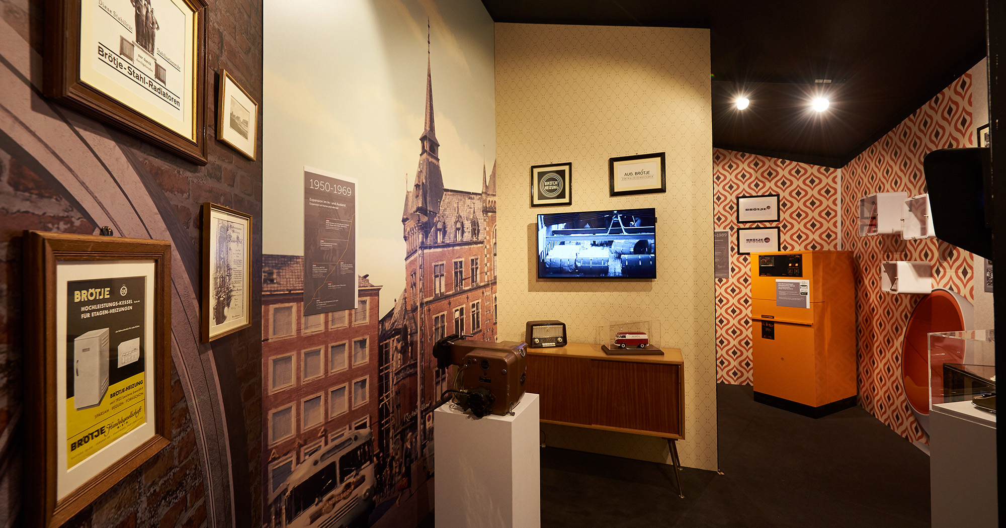Man sieht eine Kommode aus den 60ern, darauf ein altes Radio und ein Modelauto. Seitlich ist eine Wand mit einer Fototapete zu sehen, auf der ein altes Backsteingebäude abgebildet ist, darauf eine Tafel mit den Jahreszahlen „1950-1969“.