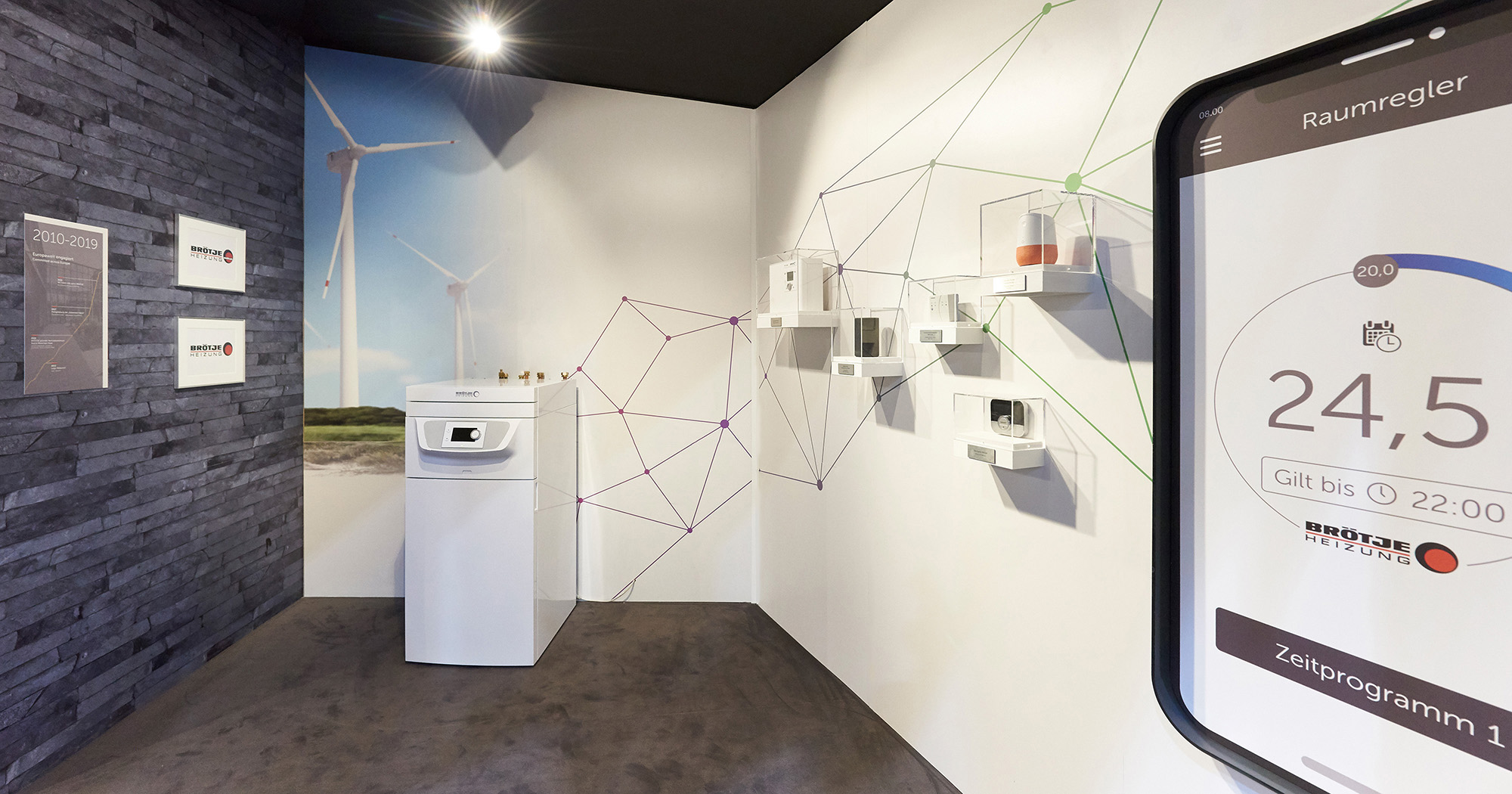 Man sieht ein modernes Ausstellungsstück von BRÖTJE, dahinter eine Fototapete, auf der Windräder zu sehen sind. Im rechten Bildrand ein riesiger Raumregler von BRÖTJE. Auf der linken Wand eine Tafel mit den Jahreszahlen „2010-2019“. 