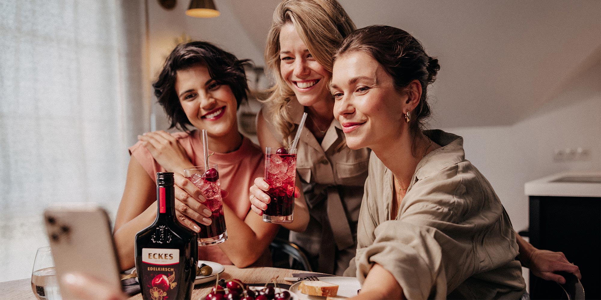 Drei Frauen machen ein Selfie und halten Cocktails aus ECKES Edelkirsch in der Hand. Eine Flasche ECKES Edelkirsch steht auf dem Tisch.