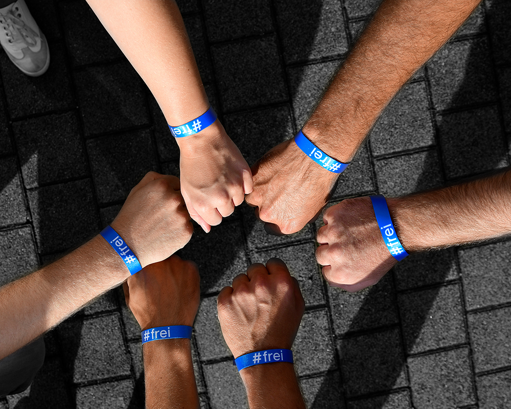 Das Foto zeigt sechs Arme, die aus dem Bildrand in die Mitte ragen. Die Hände sind zu Fäusten geballt. An den Armen erkennt man ein blaues Band auf dem in weiß „#frei“ steht.