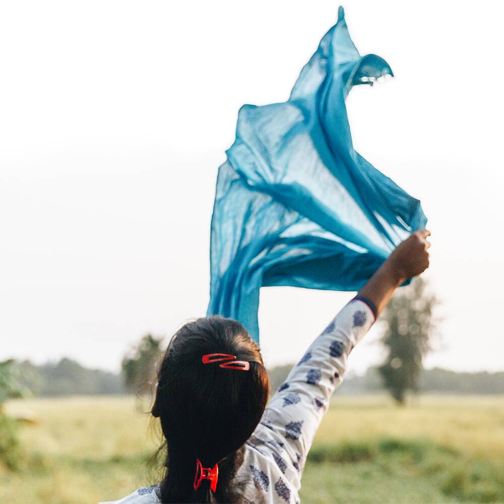 Das Foto zeigt eine Frau mit Zopf von hinten, die ein hellblaues, halbtransparentes Tuch über ihrem Kopf in den Wind hält. Im Hintergrund unscharf eine Wiese und Himmel.