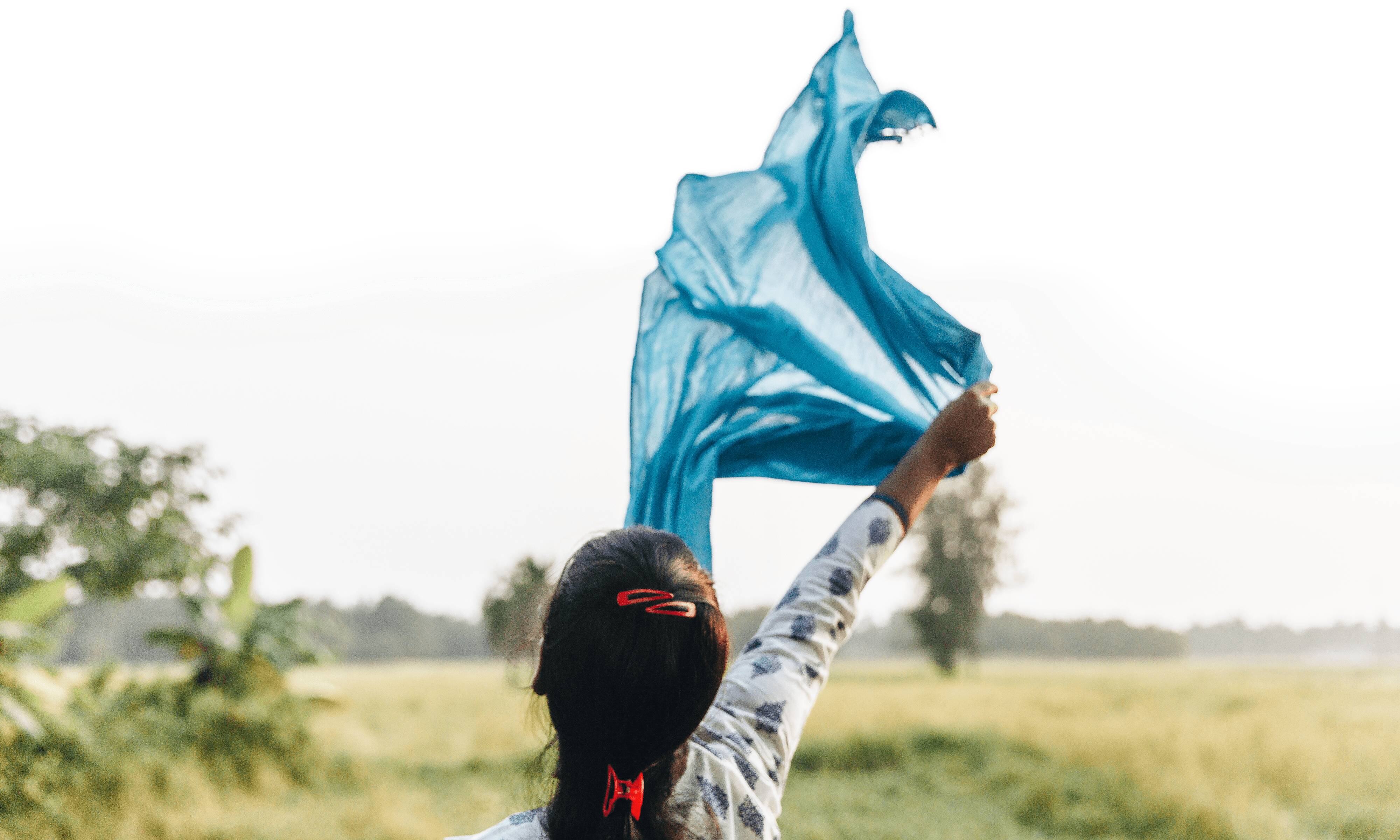 Das Foto zeigt eine Frau mit Zopf von hinten, die ein hellblaues, halbtransparentes Tuch über ihrem Kopf in den Wind hält. Im Hintergrund unscharf eine Wiese und Himmel.