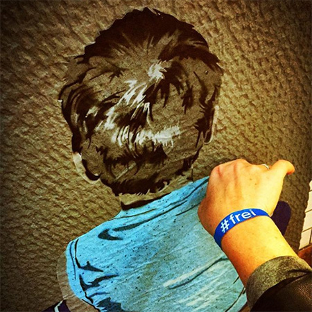 Das Foto zeigt eine Hand mit blauem #frei Band, die ins Bild ragt. Im Hintergrund eine Malerei, die einen Jungen von hinten zeigt.