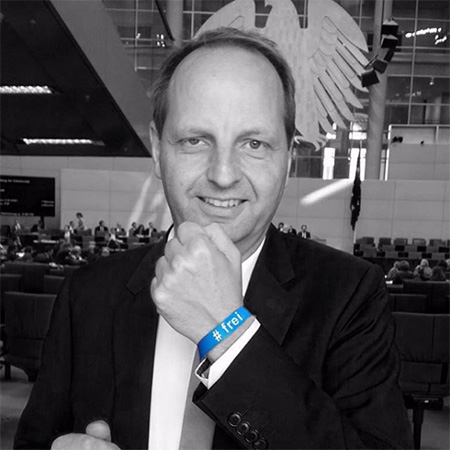 Das Foto zeigt einen Mann im Anzug vor dem Bundesadler, der seine Hand mit blauem #frei Band in die Kamera hält. Bis auf das Band ist alles schwarz/weiß.